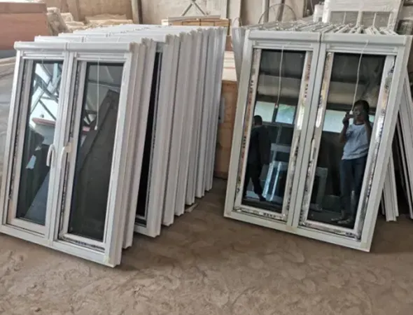 Multiple small white window frames are neatly arranged on a workbench in a busy workshop, with wooden beams and workers visible in the background.