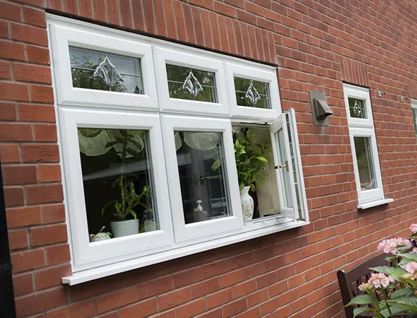 Red brick wall with white framed windows. The main window has an open pane with potted plants inside, suggesting a cozy and bright atmosphere.