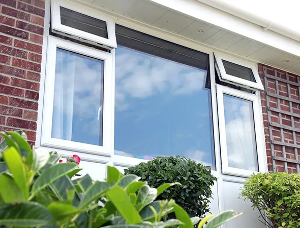 An open white-framed vinyl window set in a white wall, surrounded by lush green plants, with warm indoor light creating a peaceful contrast against the vibrant outdoor greenery.